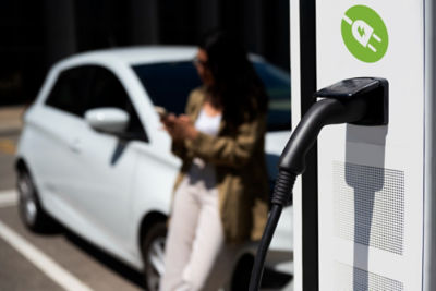 An electric vehicle (EV) charging station with a charging plug connected to the unit, while a blurred woman stands near her car in the background, raising the question: Are EV charging stations dangerous?