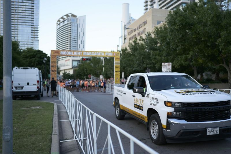 An Abacus Plumbing pickup truck parked next to barricades at the starting line of the moonlight margarita run, with participants gathered in the background and city buildings.