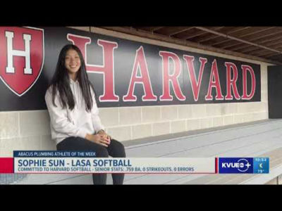 A woman sitting on a bench in front of the Harvard sign.