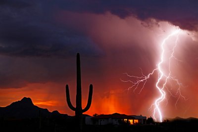 Powerful lightning strike near a house, saguaro cactus, and mountain backlit by an orange sunset and falling rain with interior lights visible in the house