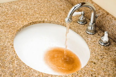 Brown dirty water pouring out of a chrome goose neck faucet into a white porcelain sink bowl in a granite vanity top