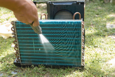 A man washes and cleans the condenser coil of an old window type air conditioning unit on the ground with a high pressure hose. Cleaning an AC unit in the yard.