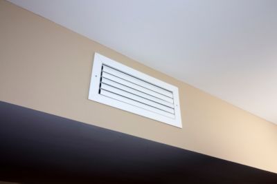 A white rectangle heating and cooling outflow air vent from a duct of an indoor HVAC system in a beige color wall near a ceiling. A small white rectangle outflow air duct vent inside a modern home.