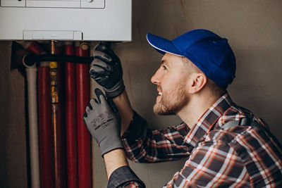 Professional Dallas plumbing technician in a blue baseball cap and plaid shirt inspecting a water heater's copper pipes while wearing protective work gloves.