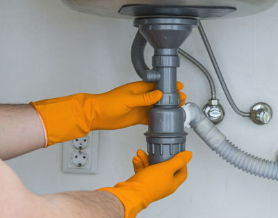 Hands with orange gloves inspecting pipes beneath sink