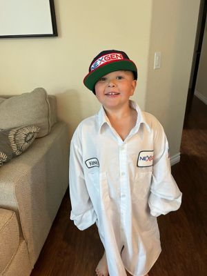 A smiling child wearing an oversized NexGen uniform shirt and hat