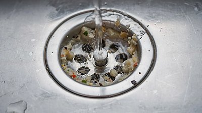 A close-up view of a kitchen sink drain shows water slowly draining through visible debris, indicating a clogged drain that may require professional help. The image highlights the potential plumbing issues caused by food waste and other particles that can lead to slow draining water and frequent blockages.