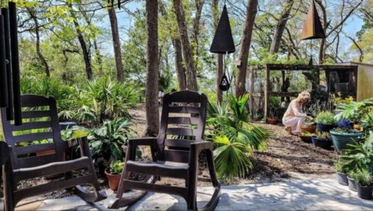  A seating are in a tree filled backyard with a woman gardening 