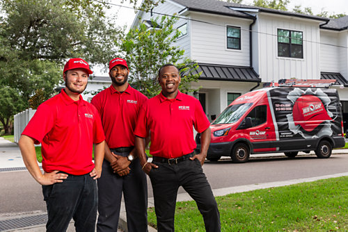 Three smiling Red Cap technicians standing in front of a company truck parked on a street in a neighborhood