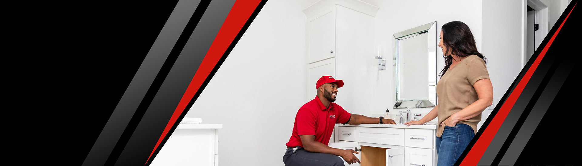 A Red Cap technician working on the plumbing of a cabinet and talking to a customer