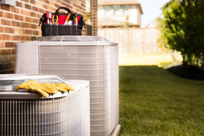 Bag of repairman's work tools, gloves on top of air conditioner units outside a brick home